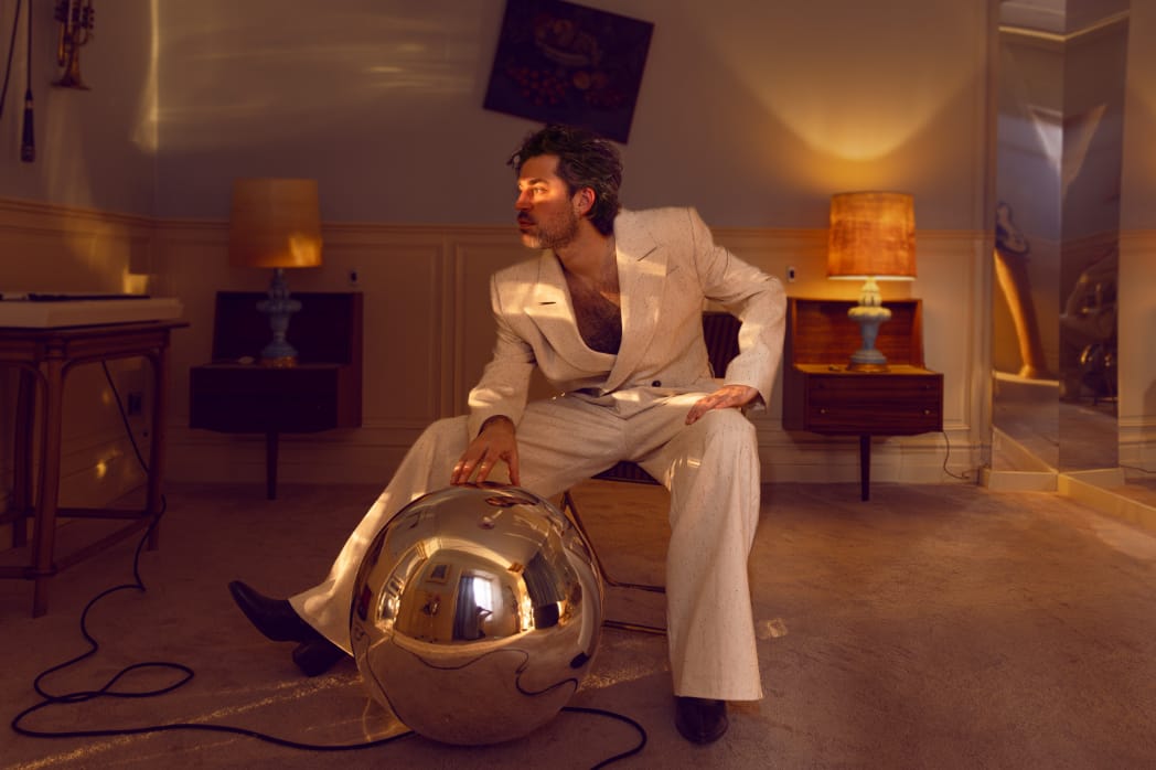 Photo of Belgium singer-songwriter J. Bernardt aka Jinte Deprez, in a white suite, posing. The vibe of the photo is very glam