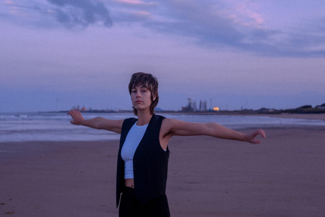 Sunset, and Newcastle-based indie-rock artist Melanie Baker standing at a beach, arms wide to the sides, looking with interes