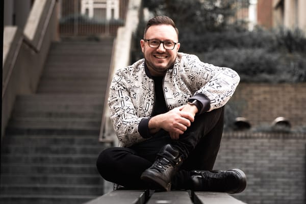 Singer, songwriter Matt Wright sitting on some stone steps, smiling, wearing black jeans and a black and white jacket.